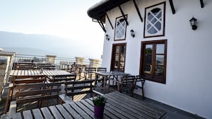 Una terraza con mesas y bancos de madera, un edificio blanco con vigas de madera oscura, y vistas a montañas distantes.