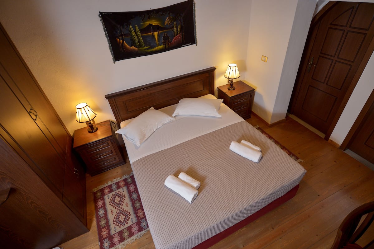 A neatly made bed with white linens, two bedside tables with lamps, a wooden door, and a painting on the wall.