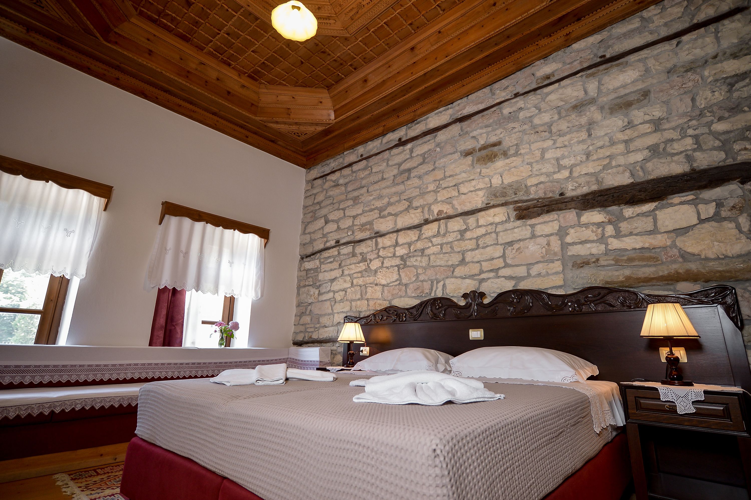 A hotel room with a stone wall, wooden ceiling, a large bed with white linens, two bedside lamps, and a window with curtains.