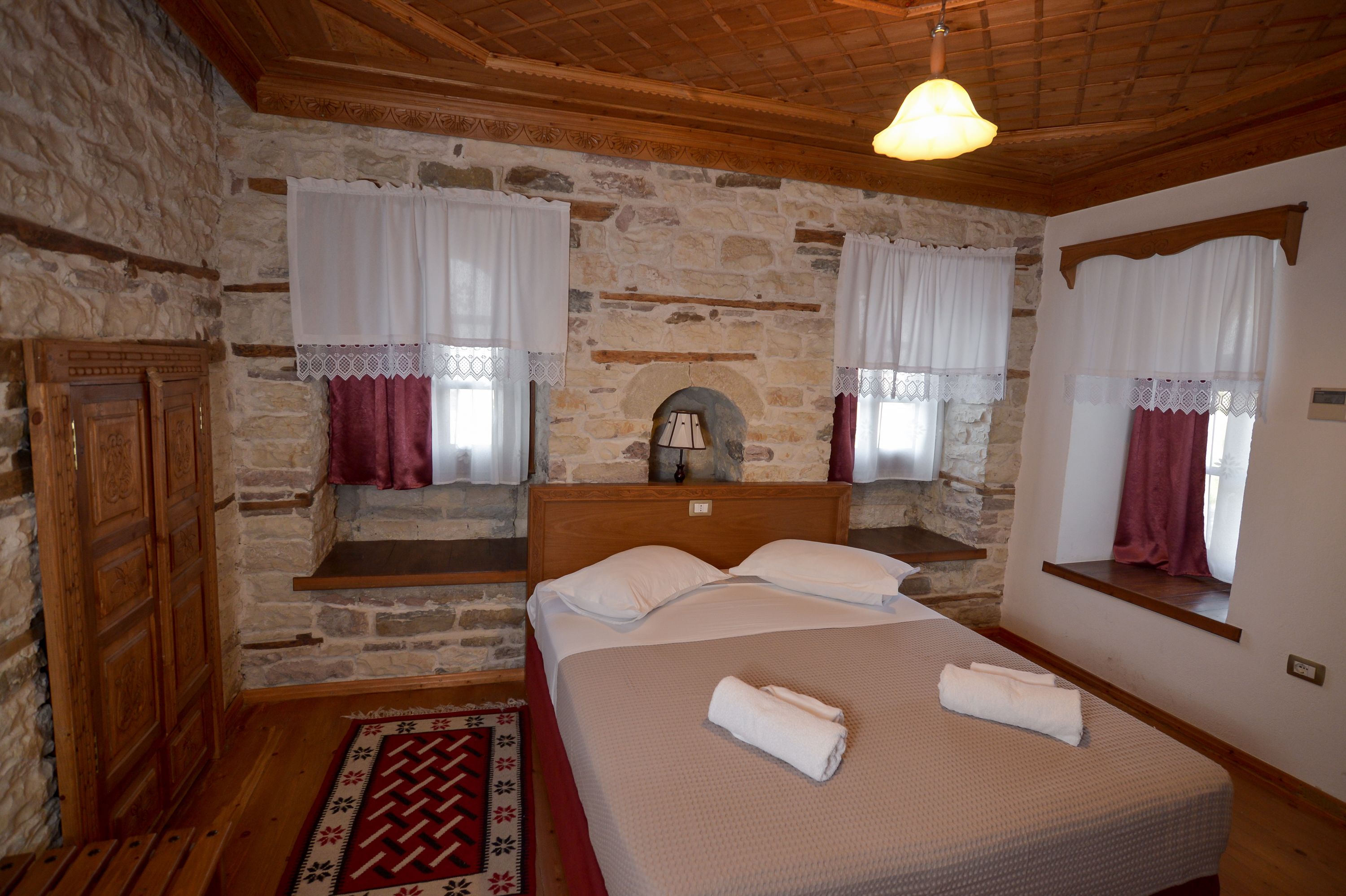 A bedroom with stone walls, a wooden ceiling, a bed with white linens, and a red patterned rug.