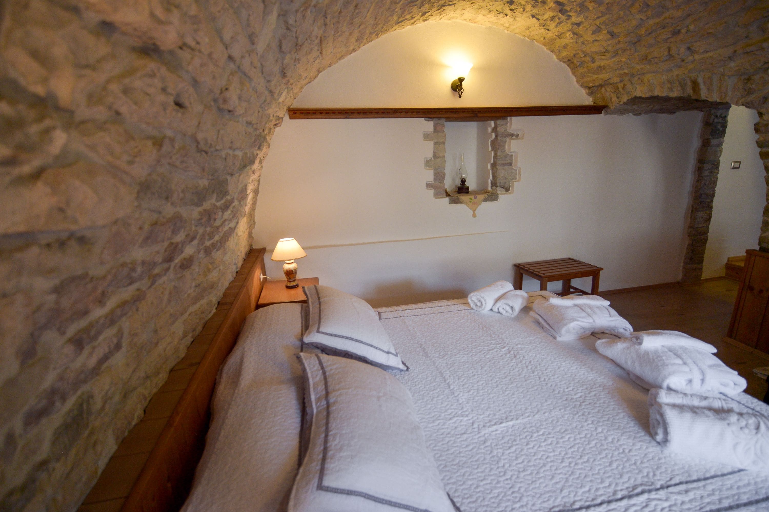 Un dormitorio con pared de piedra, una cama con ropa blanca, una mesita de noche con lámpara y una pequeña mesa de madera.