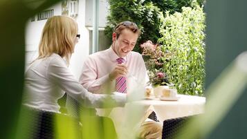 A couple dining at an outdoor table with a view of greenery and a building in the background.