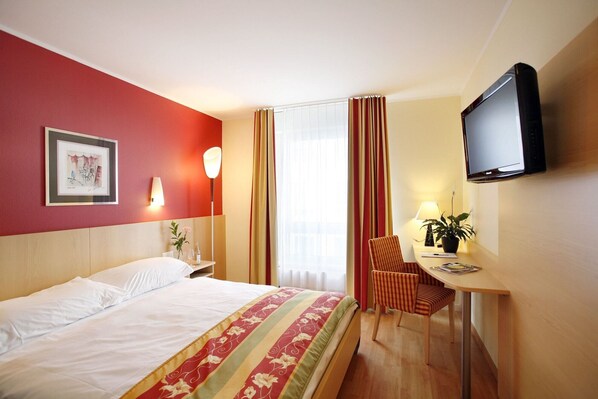 A hotel room with a bed, a desk with a chair, a television, and a window with curtains. - Ambassador (Lucerne)