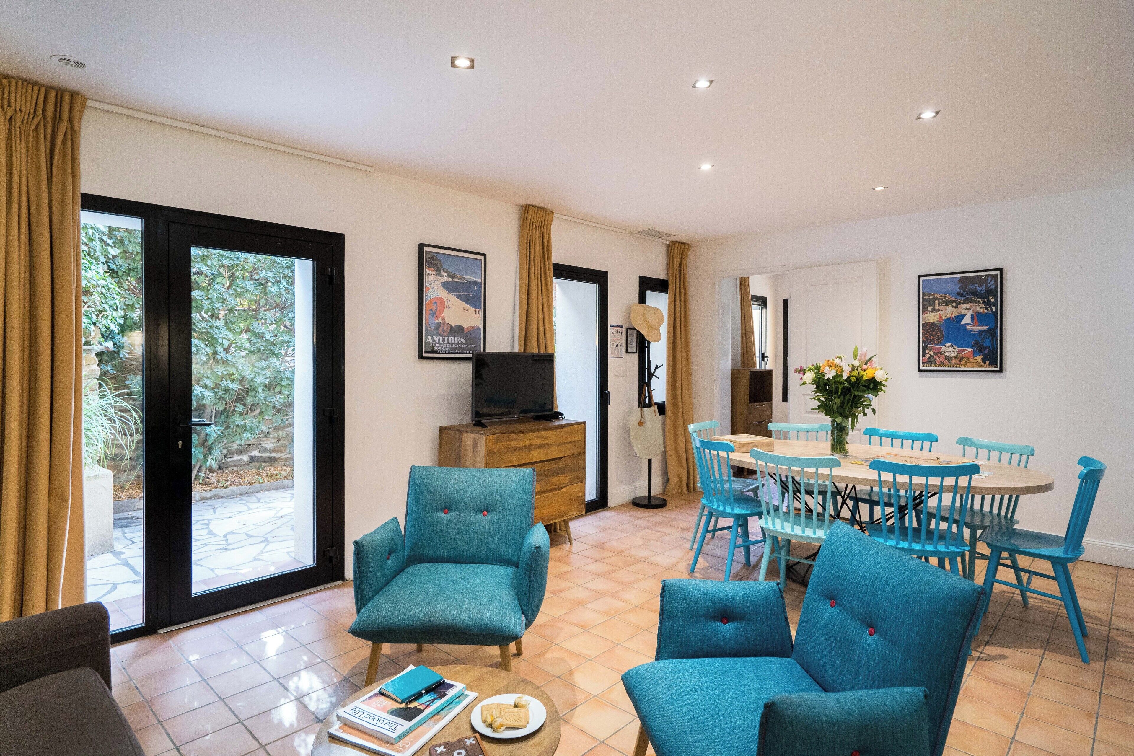 Un vaste espace de vie avec une grande porte-fenêtre coulissante donnant sur un patio, un coin repas avec des chaises bleues et un téléviseur posé sur un meuble en bois.