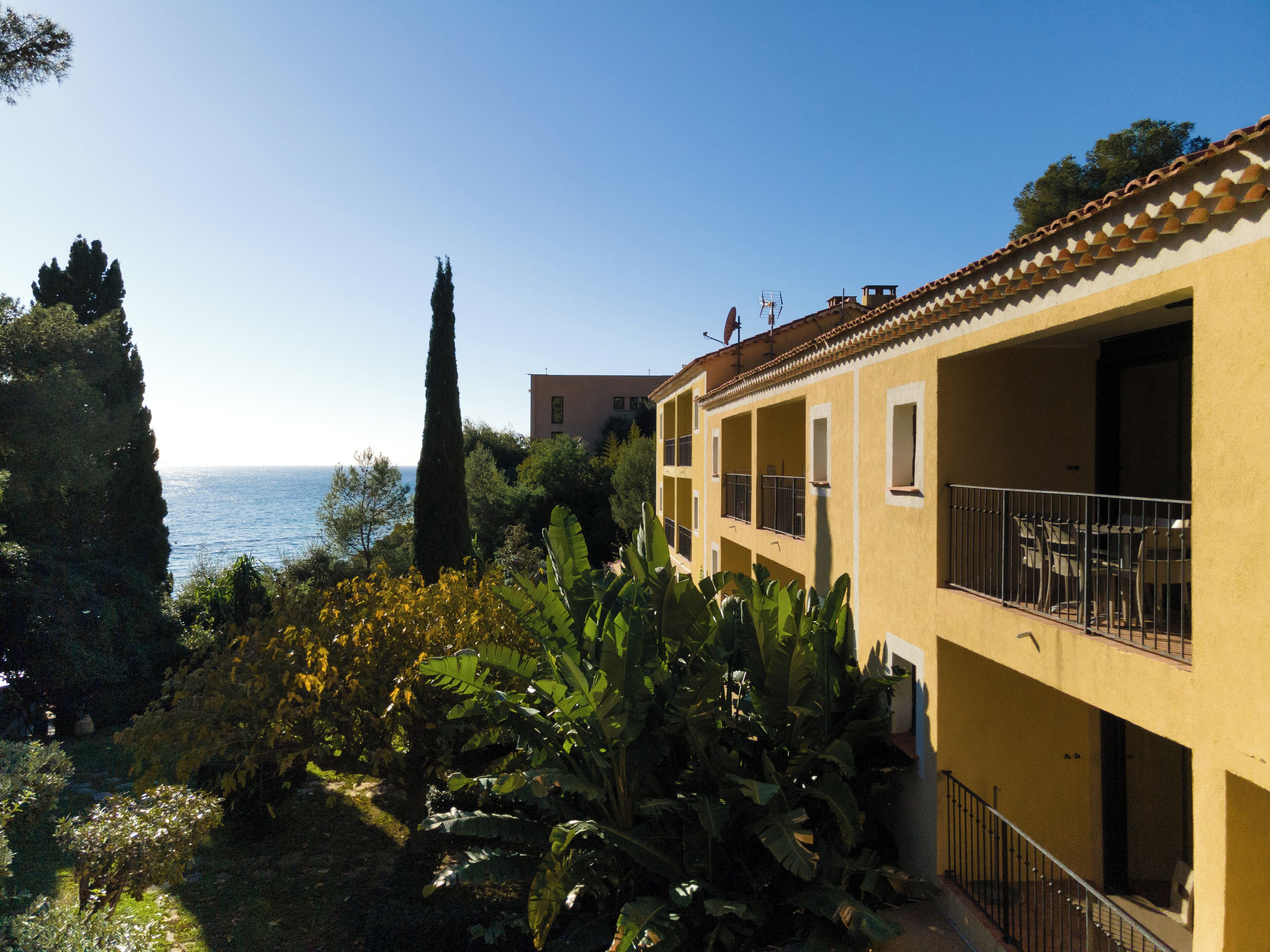 A coastal building with balconies overlooking the sea, surrounded by greenery.