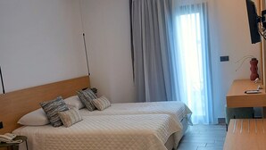 A hotel room with two beds, a wooden headboard, a small table with a telephone, a chair, and a desk with a plant. - Bianco Hotel (Leros)
