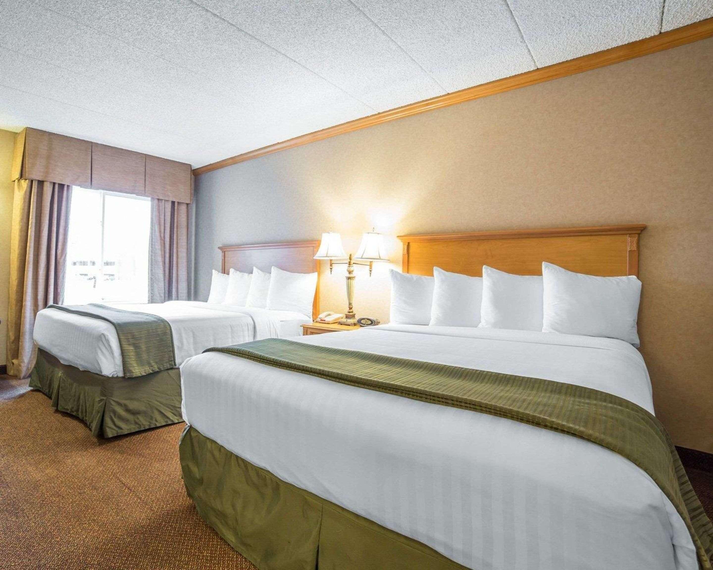 A hotel room with two beds, a wooden headboard, a nightstand with a lamp, and a window with curtains.