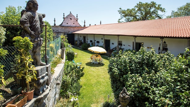 A courtyard with a statue, a building with a red-tiled roof, and a table with chairs under an umbrella. - Quinta De Santa Baia (Ponte de Lima)