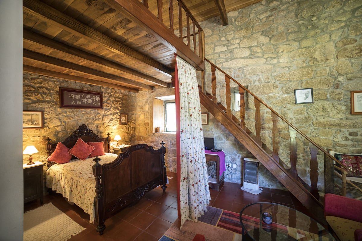 A bedroom with a wooden bed, stone walls, a wooden staircase, and a stone fireplace.