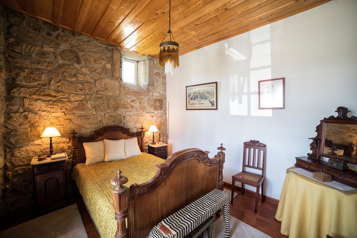 A bedroom with a stone wall, wooden ceiling, a bed with a wooden headboard, two bedside tables with lamps, a chair, a bench, and a table with a mirror and picture frames.