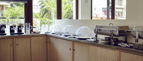 A kitchen with wooden cabinets, a coffee machine, a milk dispenser, and a buffet setup.