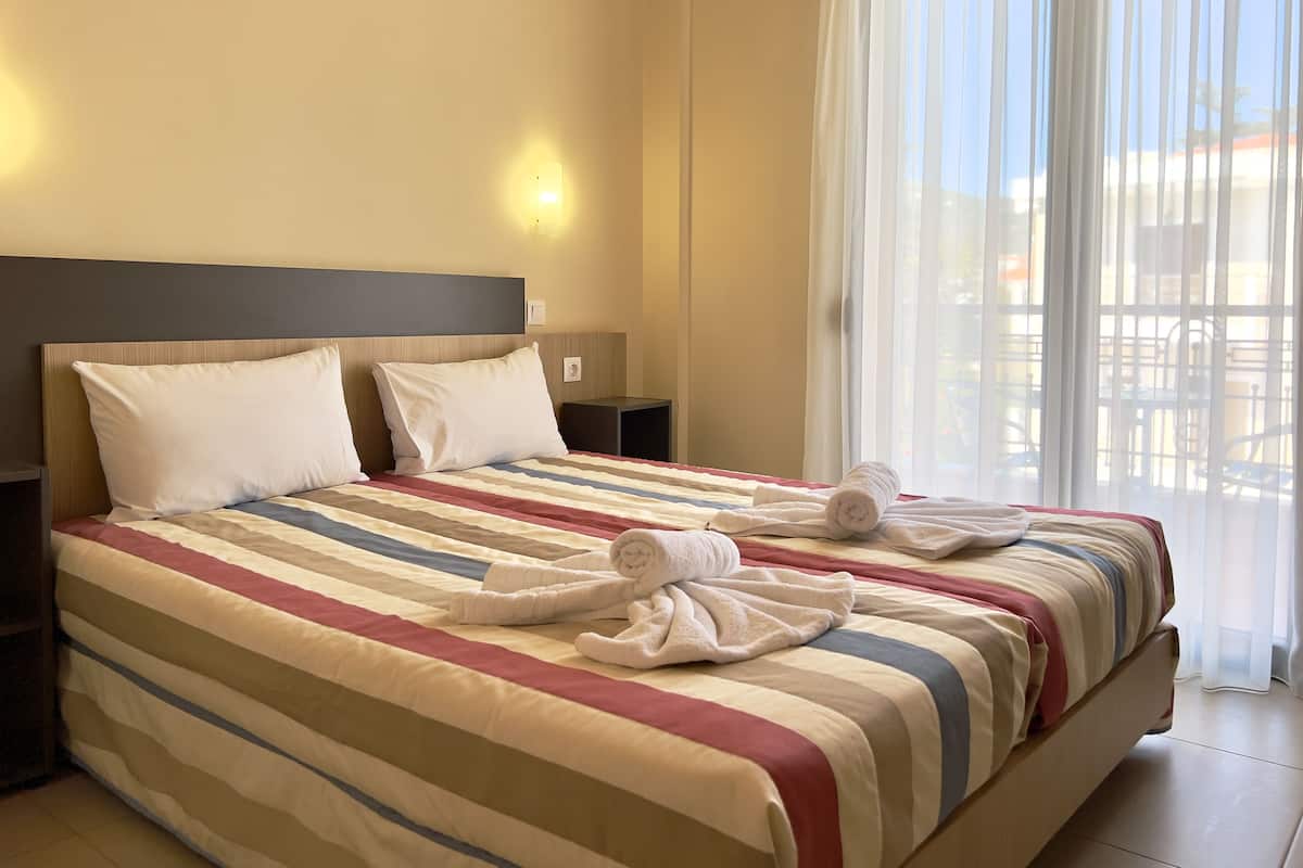 A hotel room with a large bed, two pillows, a striped bedspread, and a window with sheer curtains.