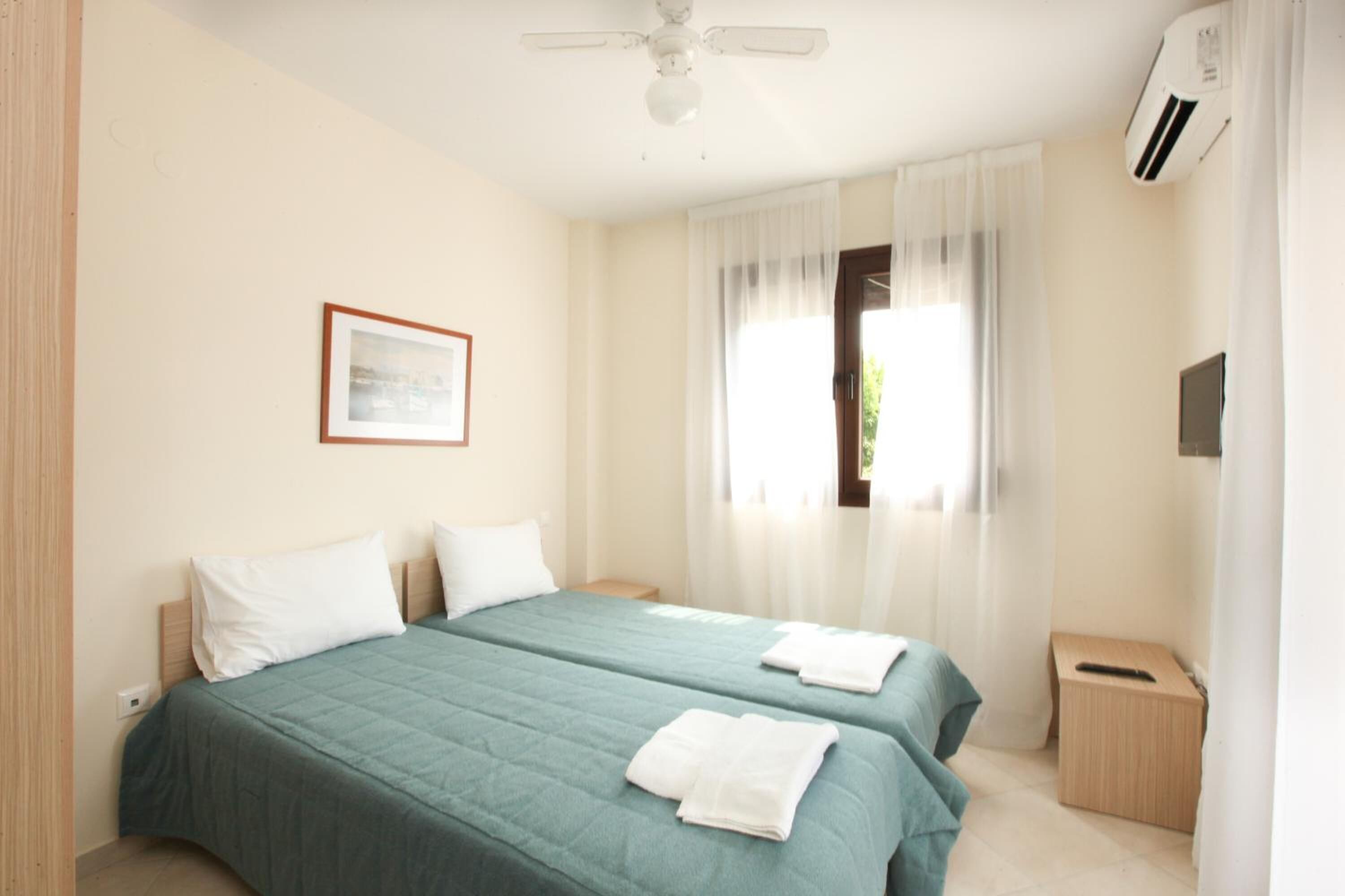 A hotel room with two beds, a ceiling fan, an air conditioning unit, a framed picture on the wall, and a window with sheer curtains.
