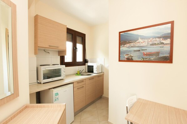 A compact kitchen with wooden cabinets, a microwave, a toaster, and a sink. There's a small table and a chair, and a framed picture of a coastal town on the wall. - Skiathos Somnia (Skiathos)