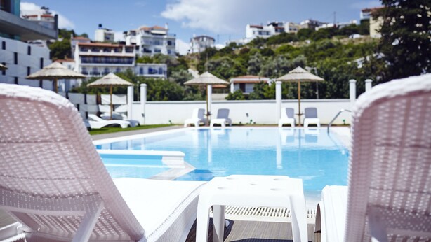 Zona de piscina amb gandules i ombrel·les, una piscina blava cristal·lina i edificis al fons.