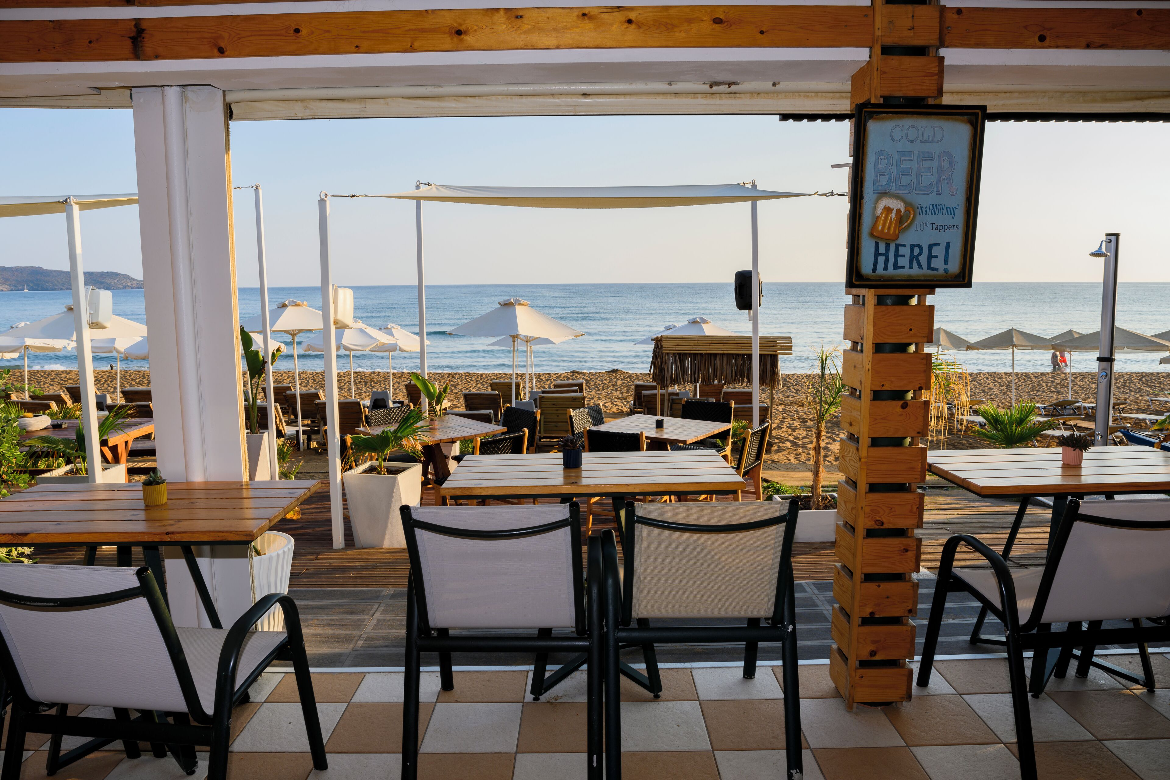 Un espace extérieur aménagé avec des tables et des chaises, donnant sur une plage parsemée de parasols et où est affiché un panneau publicitaire pour de la bière fraîche.