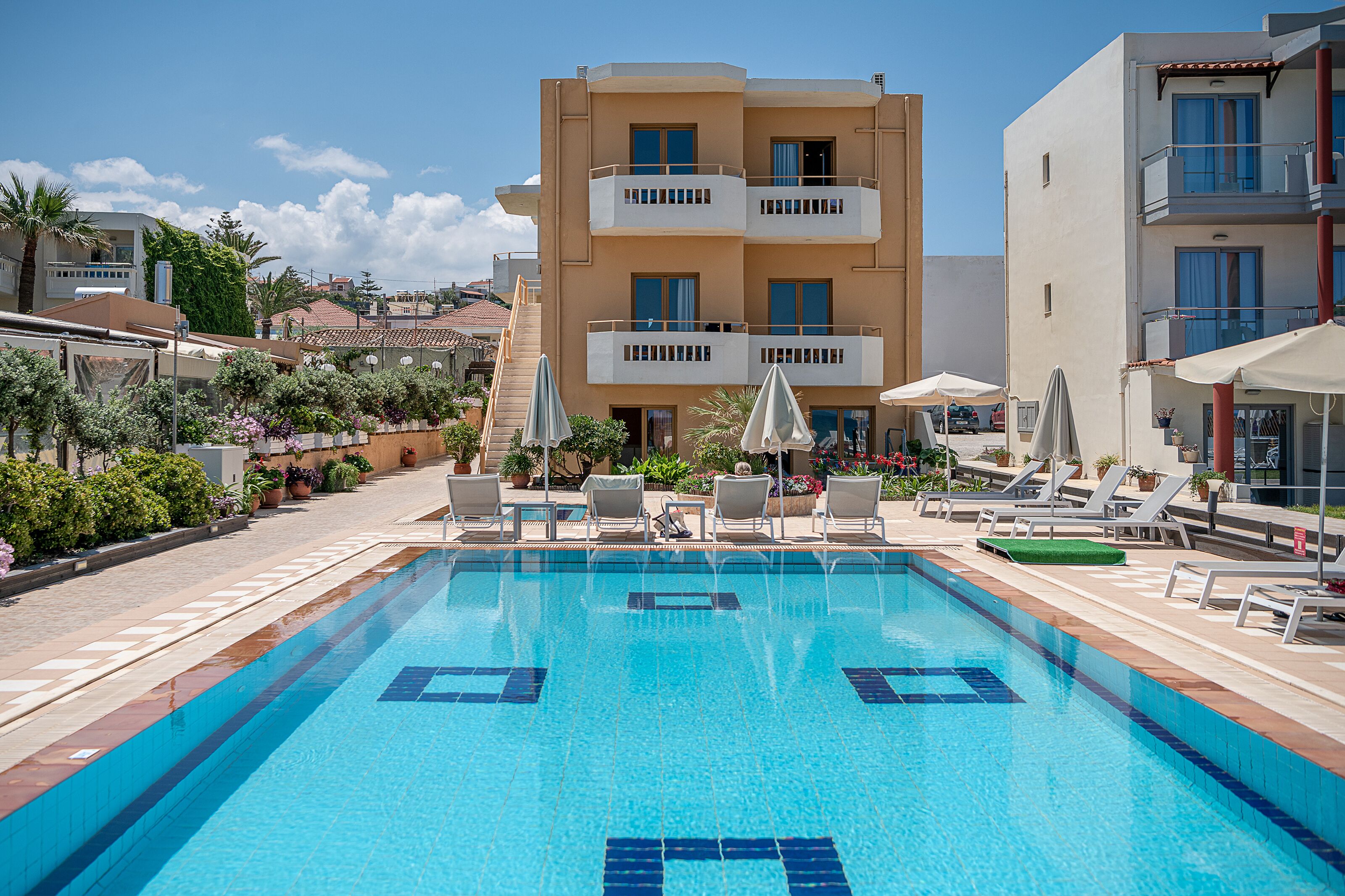 A swimming pool with lounge chairs and umbrellas, surrounded by buildings with balconies and a clear sky.