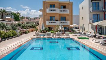 A swimming pool with lounge chairs and umbrellas, surrounded by buildings with balconies and a clear sky.