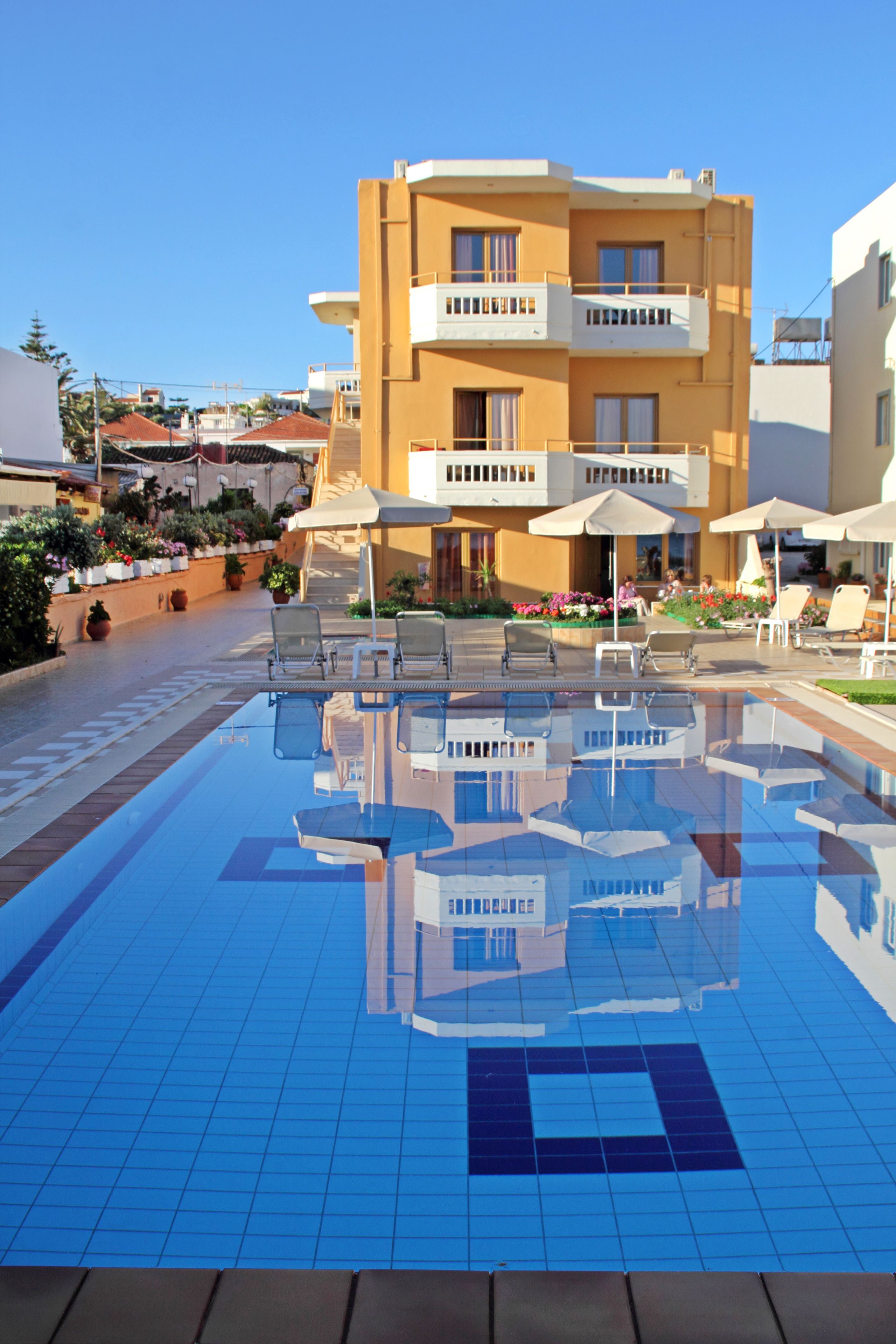 A swimming pool with lounge chairs and umbrellas, surrounded by a building with balconies and a garden.