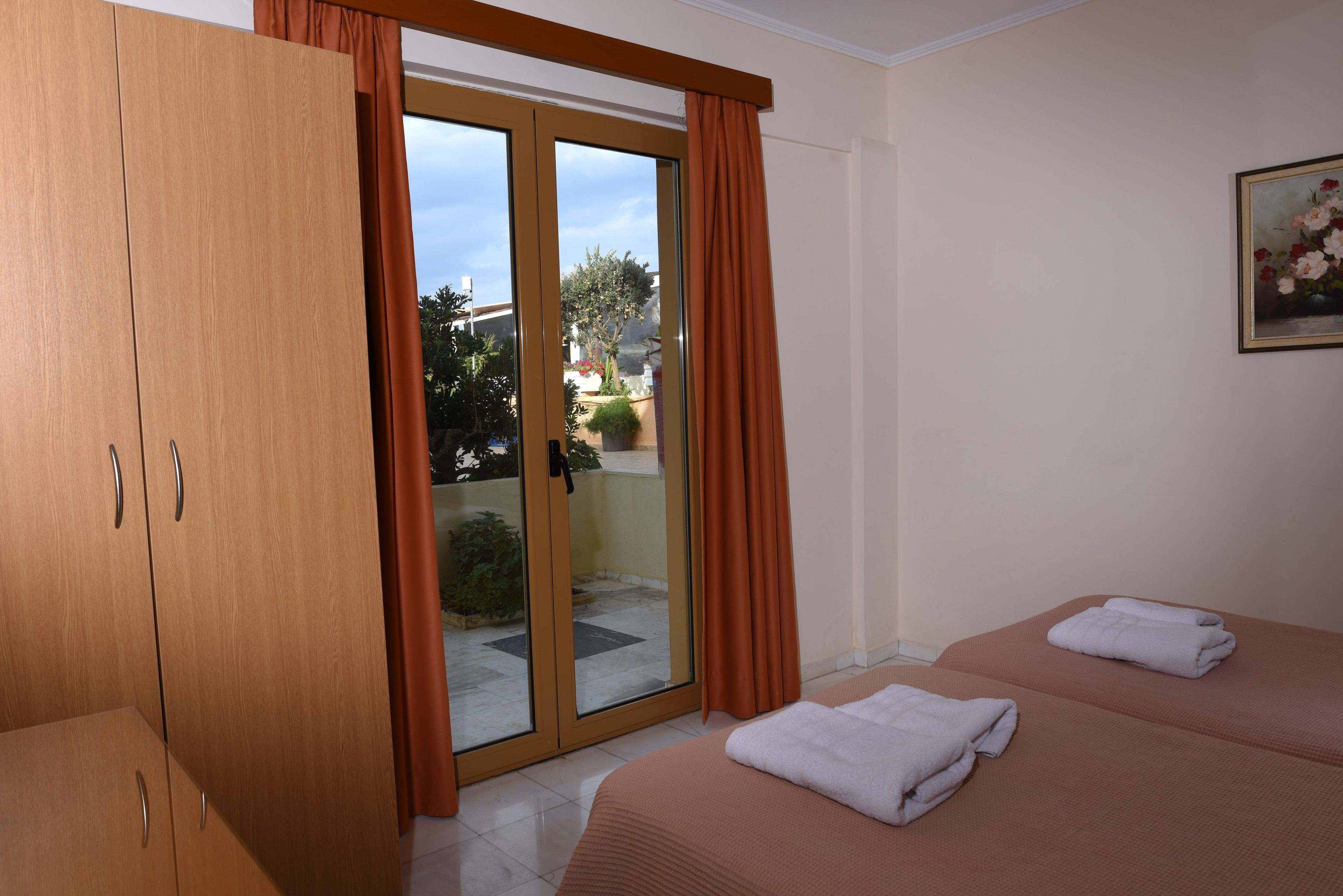 Habitación de hotel con puerta corredera de vidrio a un balcón, cama con cubierta color beige y armario de madera.