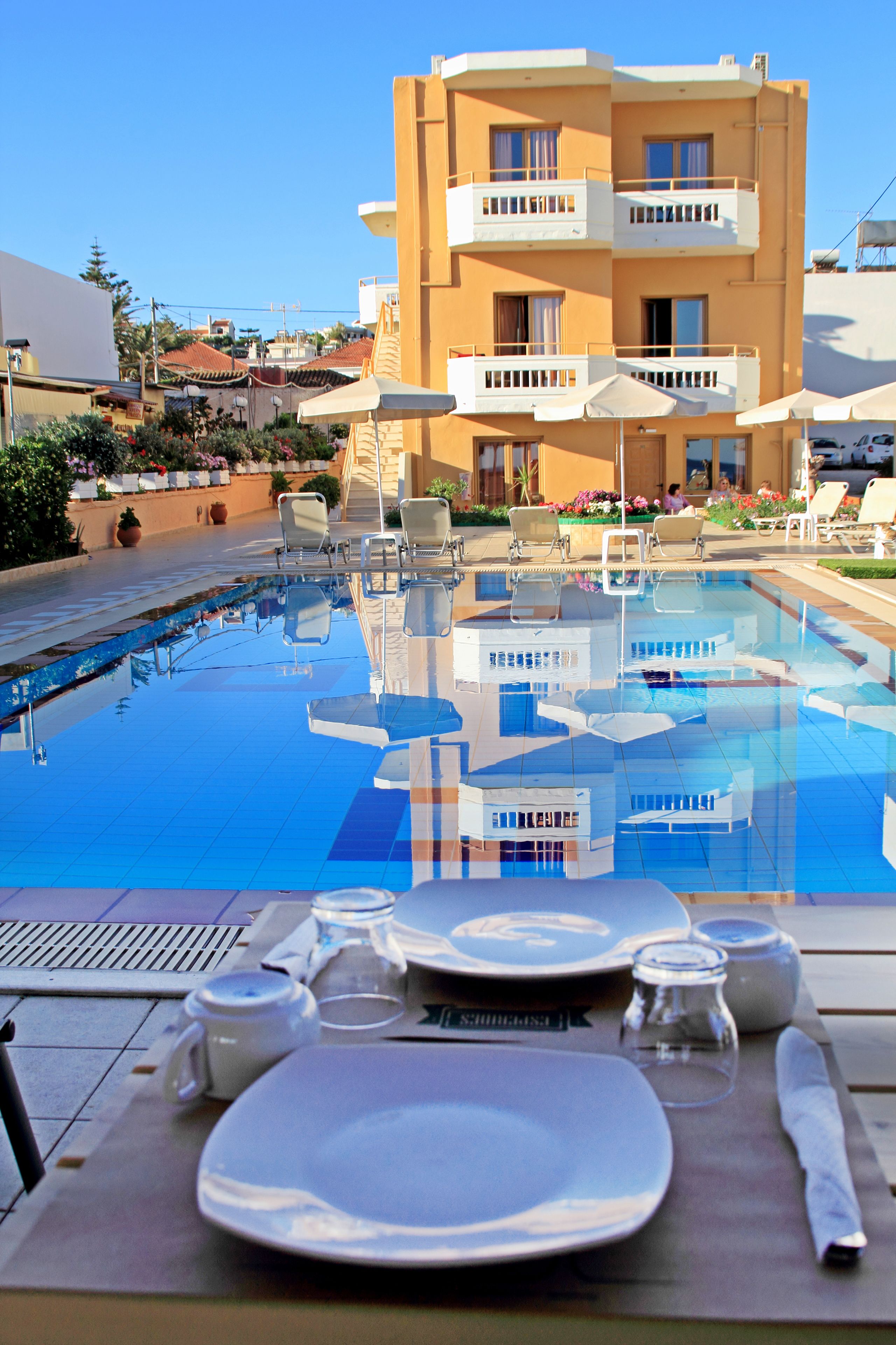 Ein Essbereich am Pool mit einem Tisch für zwei Personen, freie Sicht auf den Pool und ein Gebäude mit Balkonen im Hintergrund.