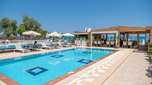 Un espace piscine avec des chaises longues et des parasols, un coin salon couvert et une vue sur la mer.