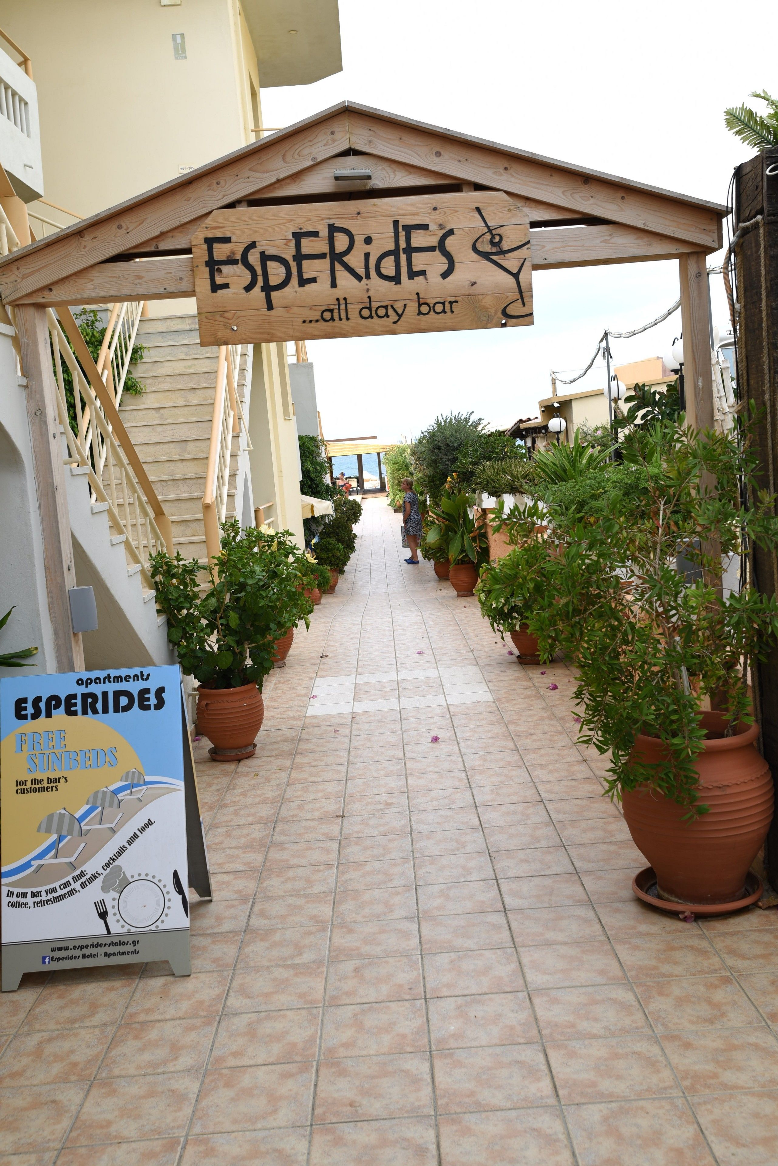 Un sendero bordeado de macetas con plantas que lleva a una pérgola de madera con un letrero que dice 'Esperides all day bar'.