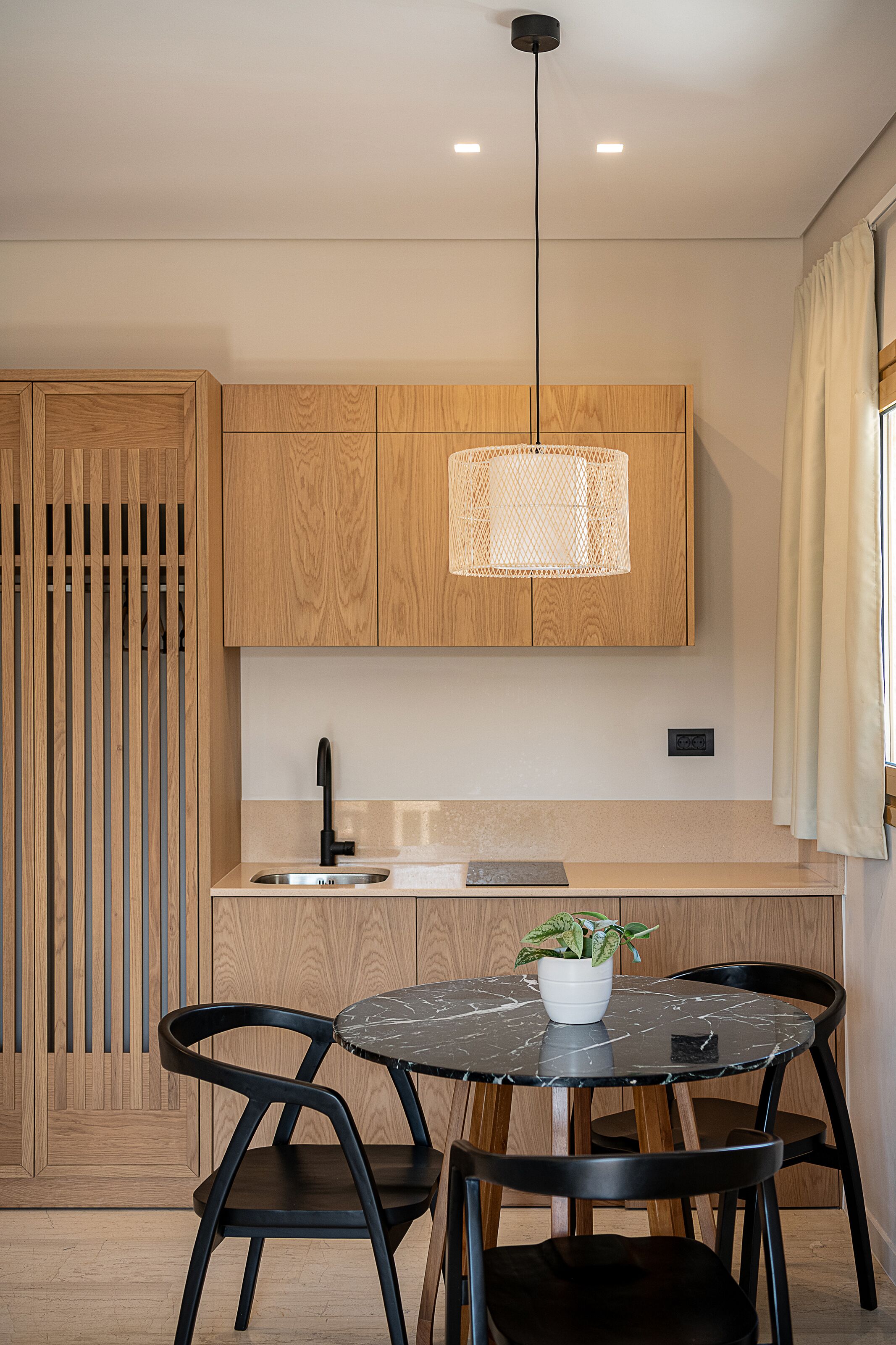 A modern kitchen with wooden cabinets, a black faucet, a round marble table with two black chairs, and a hanging light fixture.