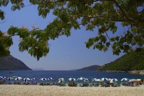 Ein Strand mit Sonnenschirmen und Menschen, ein klarer Himmel und eine hügelige Landschaft im Hintergrund.