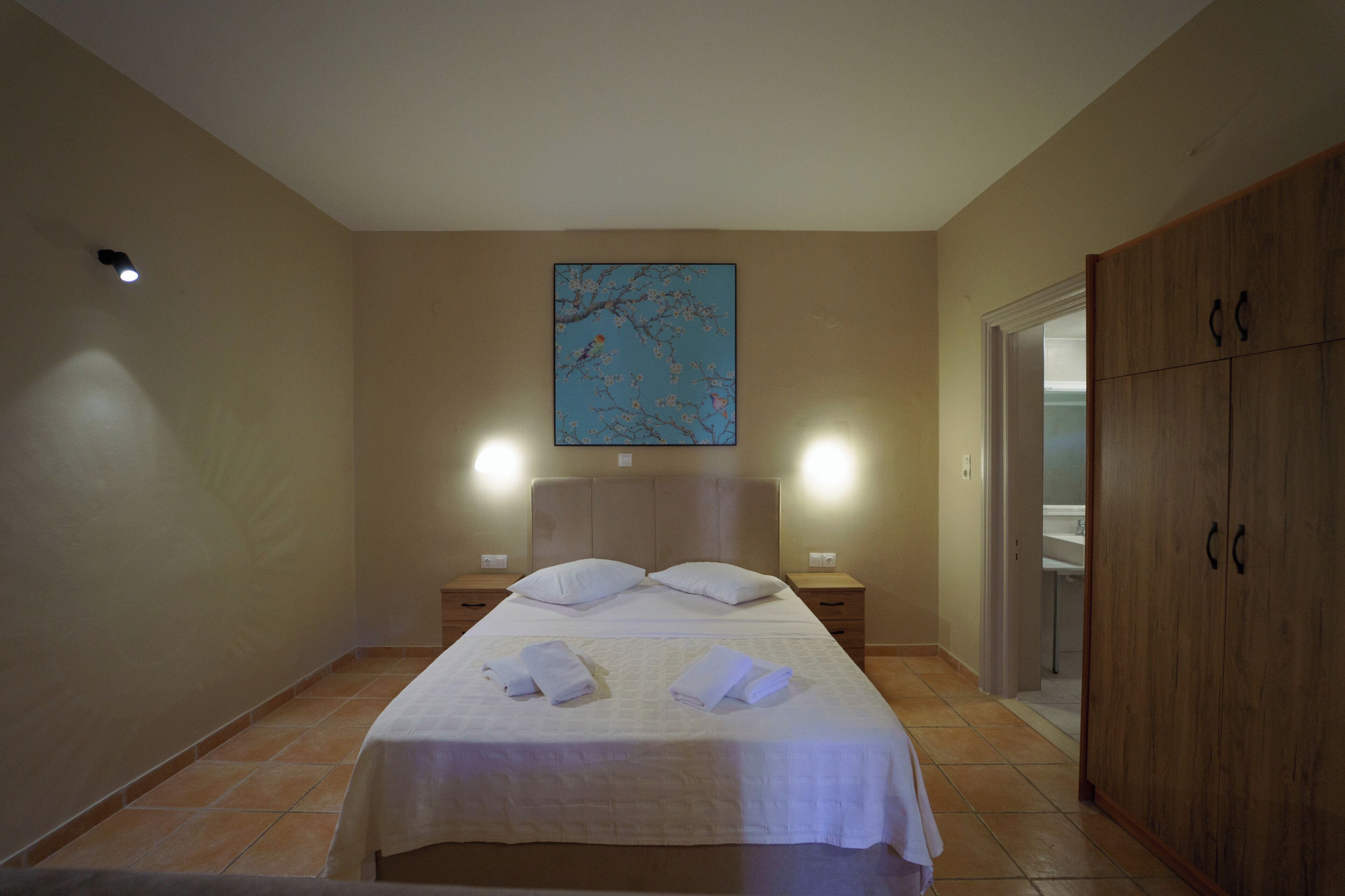 A hotel room with a bed, two bedside tables, a nightstand, a wardrobe, and a painting on the wall.