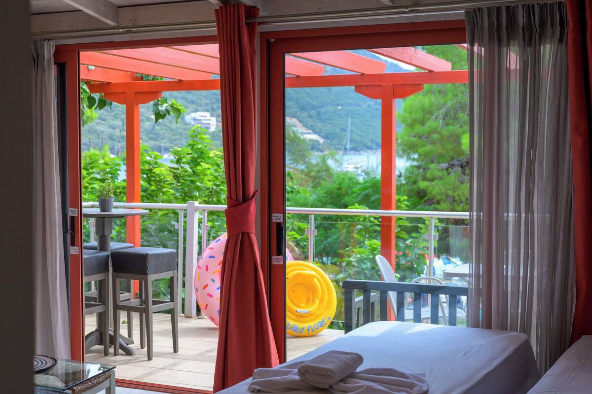 A hotel room with a balcony, a bed, a table with chairs, and a view of greenery and a body of water.