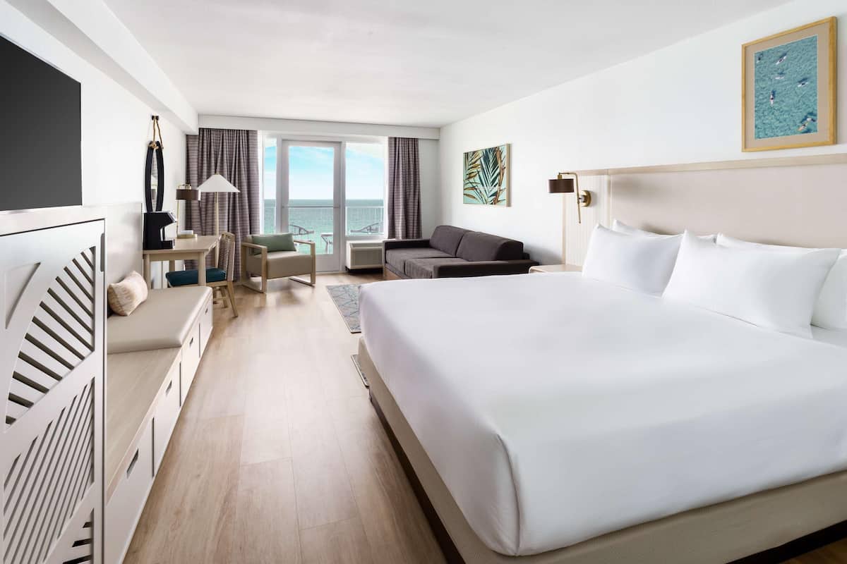 Een hotelkamer met een groot bed, een bureau, een bank, een televisie en uitzicht op zee door de ramen.