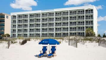 A multi-story hotel with a white facade and balconies. There are beach chairs and a blue umbrella in the foreground.