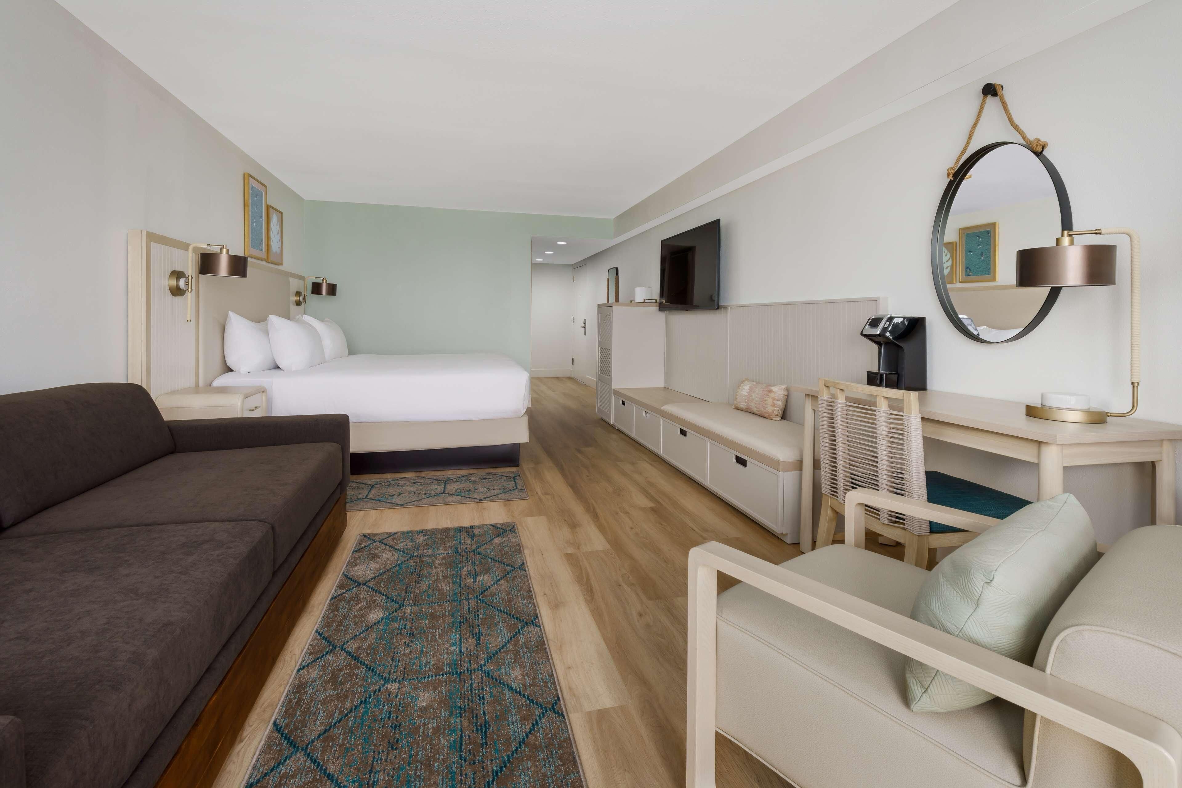 A hotel room with a bed, a sofa, a desk with a chair, a TV, and a mirror.