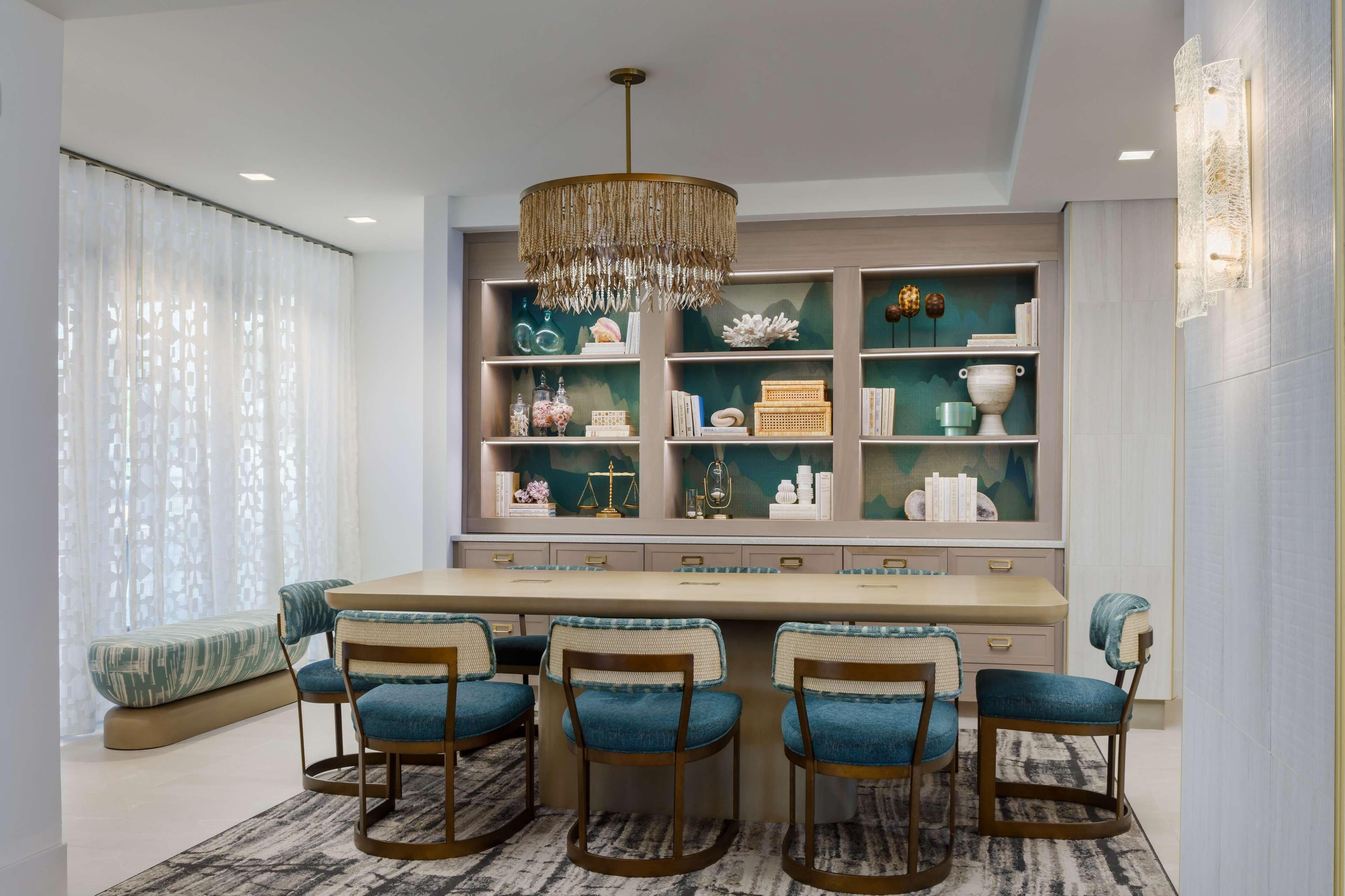 A dining area with a long table, four bar stools, and a chandelier. Shelves with decorative items are behind the table.