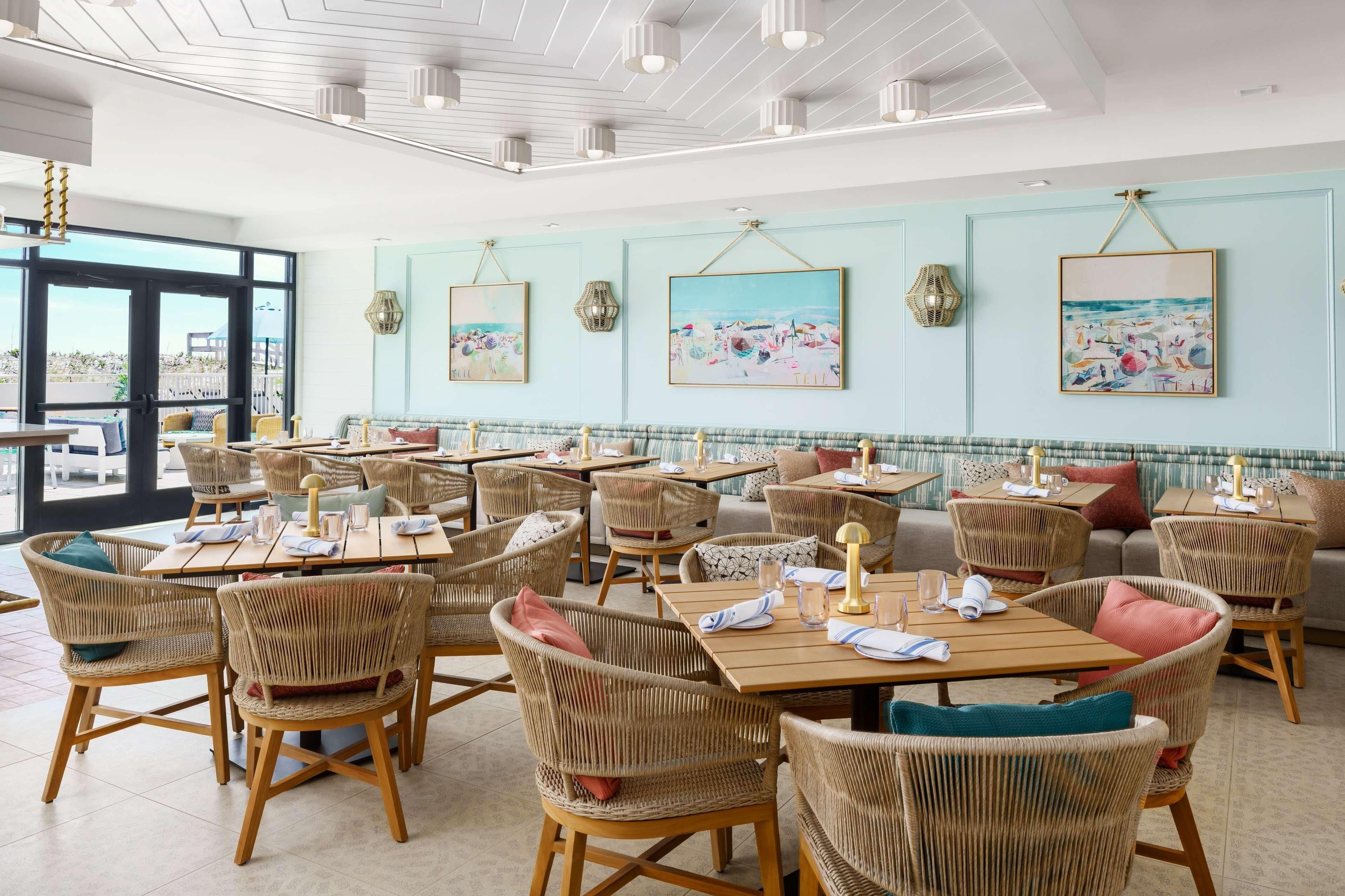 A restaurant with wicker chairs and tables, framed artwork on the walls, and a view through large windows.