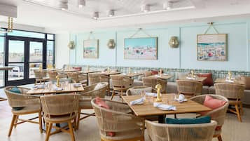 A restaurant with wicker chairs and tables, framed artwork on the walls, and a view through large windows.