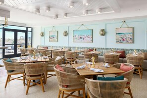 Un restaurant avec des chaises et des tables en osier, des œuvres d’art encadrées aux murs et une vue dégagée grâce à de grandes fenêtres.