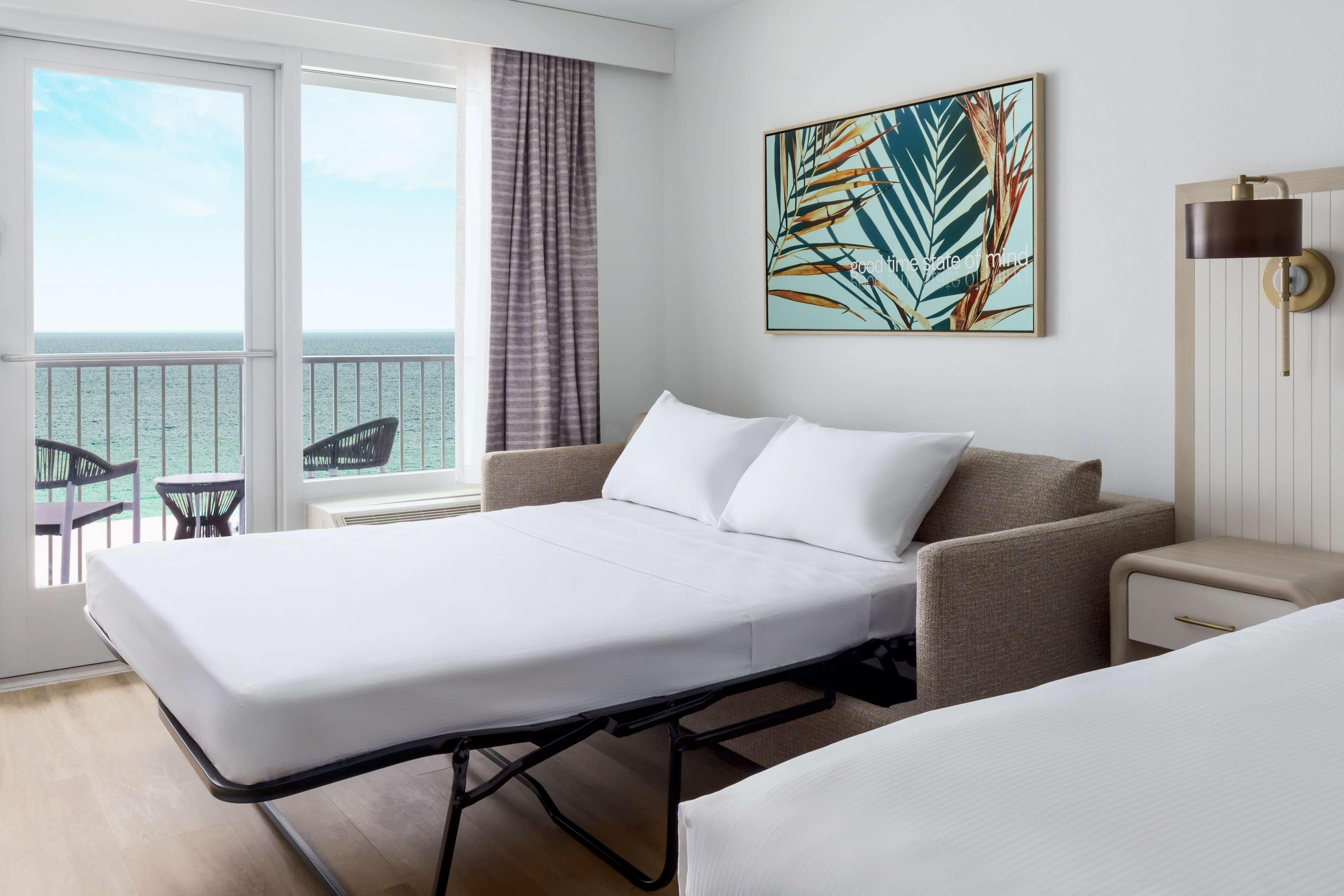 A hotel room with a bed, a sofa, a small table, a chair, and a view of the sea through large windows.