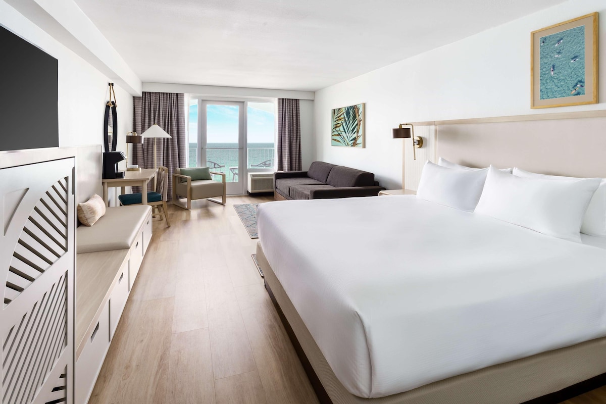 Una camera d'albergo con un letto grande, una scrivania, un divano, una TV e vista mare dalle finestre.