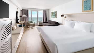 A hotel room with a large bed, a desk, a sofa, a TV, and a view of the sea through the windows.