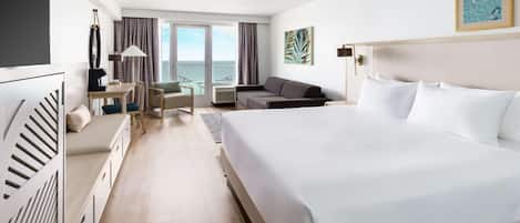 A hotel room with a large bed, a desk, a sofa, a TV, and a view of the sea through the windows.