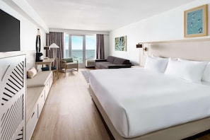 A hotel room with a large bed, a desk, a sofa, a TV, and a view of the sea through the windows.