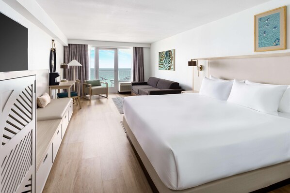 Une chambre d’hôtel avec un grand lit, un bureau, un canapé, une télévision et une vue sur la mer par les fenêtres.
