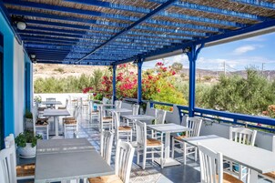 Un patio exterior con mesas y sillas blancas, techo azul y vistas del paisaje.
