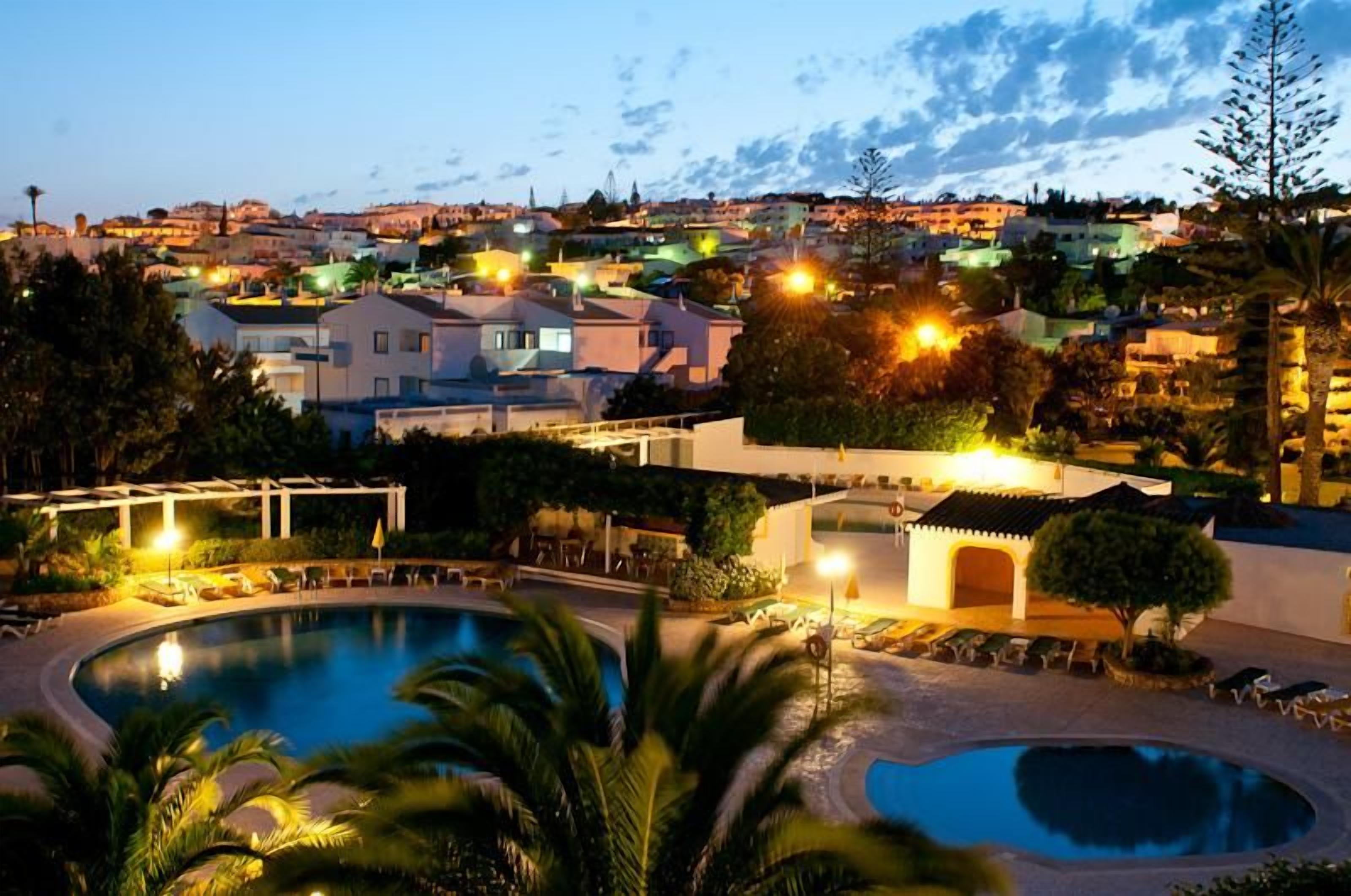 Un resort con zona de piscina, sillones de descanso y vistas del pueblo al anochecer.