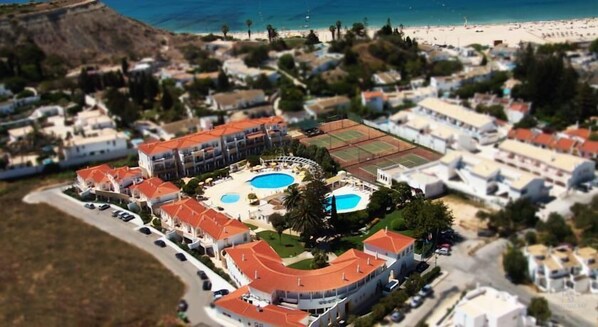 Vista aérea de un complejo turístico con varias edificaciones, un área de piscina central y una playa al fondo.