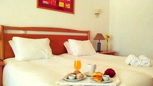 Una cama bien tendida con sábanas blancas, una bandeja con desayuno que incluye cruasanes, una taza de café y zumo de naranja.