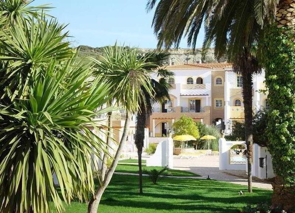Un immeuble à plusieurs étages doté de balcons, entouré d’une végétation luxuriante et de palmiers.