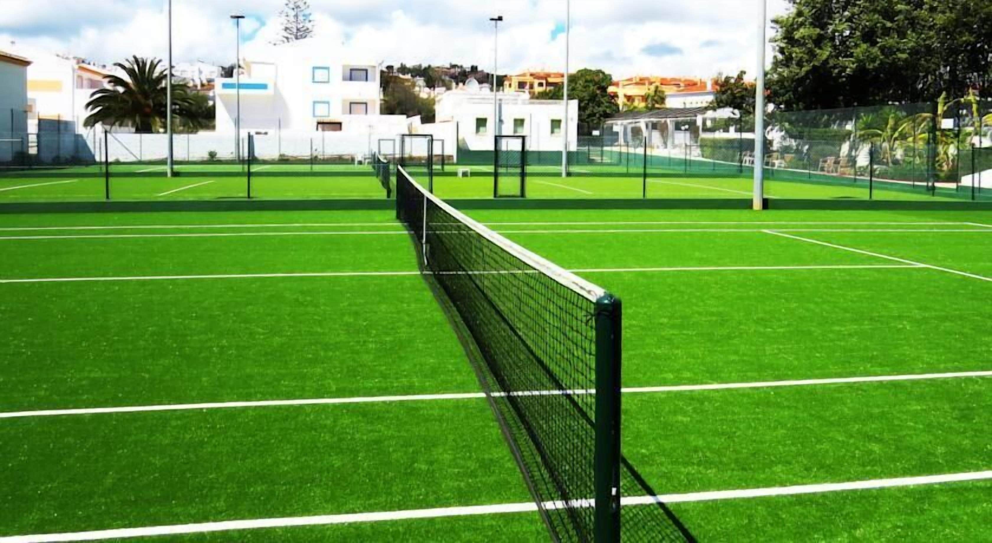 A tennis court with a net in the center, surrounded by a fence and buildings in the background.